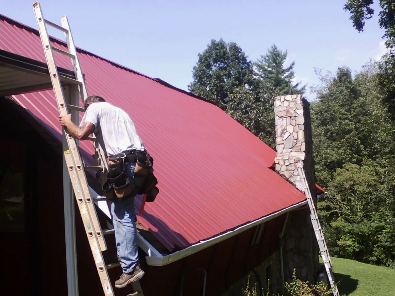 Roofer on a ladder beside a red metal roof and stone chimney — Soffit & Fascia Repair by Roofing Brothers in Bagley